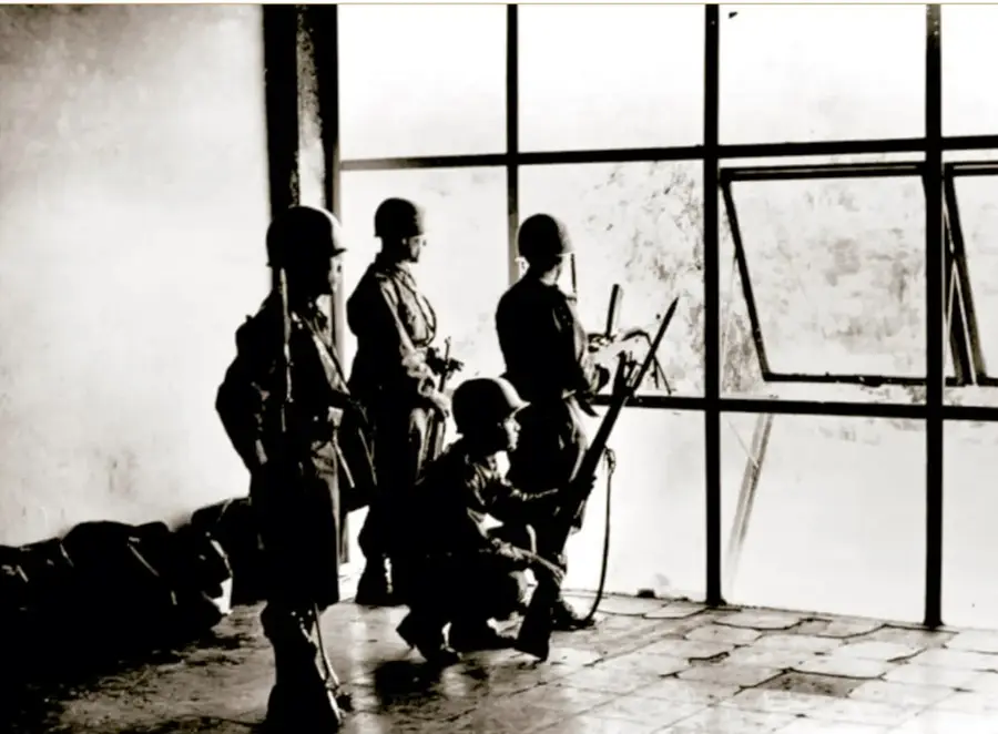 Fotografía en blanco y negro de cuatro granaderos armados con cascos, en la ventana de un departamento de la Ciudad de México en 1968; uno está en cuclillas y los demás de pie, vigilando el exterior a través de grandes ventanales. Pie: INAH, Secretaría de Cultura, SINAFO.FN.MX.