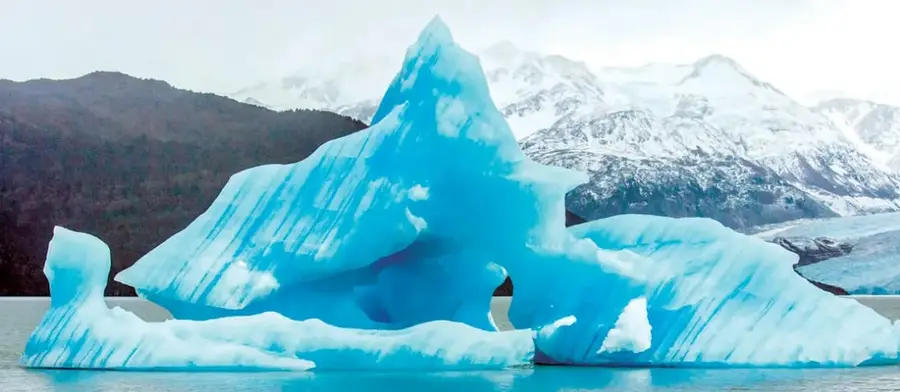 Fotografía de un gran bloque de hielo azul (témpano) del Glaciar Grey, flotando en un lago, con montañas nevadas y bosque al fondo. Corresponde a la Patagonia chilena.