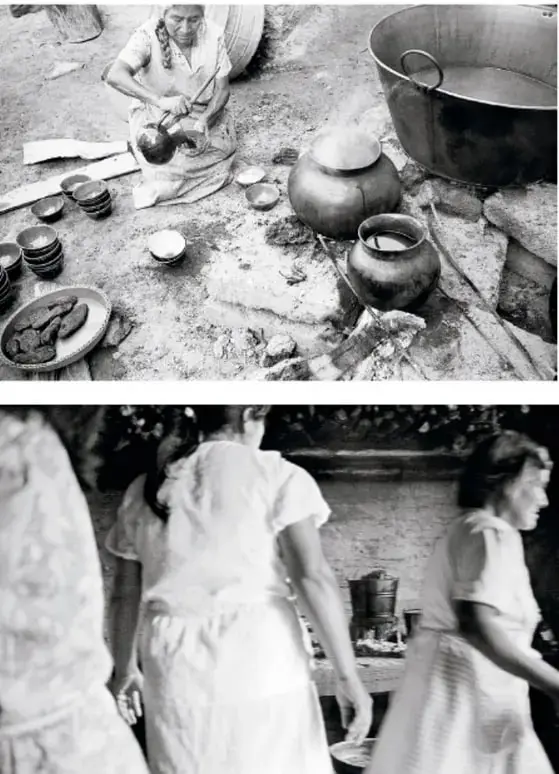 Fotografía en blanco y negro de una mujer sentada en el suelo preparando alimentos rodeada de ollas de barro, cazos grandes, platos y utensilios de cocina.