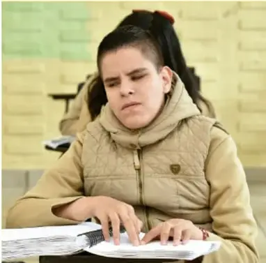 Fotografía de un estudiante con moño y chamarrón beige que lee con los dedos un libro grueso de páginas en braille, sentado en un salón de clases.