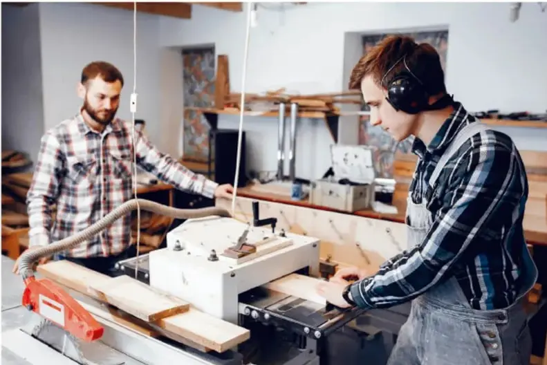 Fotografía de dos personas trabajando en un taller de carpintería; una opera una máquina para procesar tablas de madera mientras la otra observa, ambos con equipo de protección auditiva.