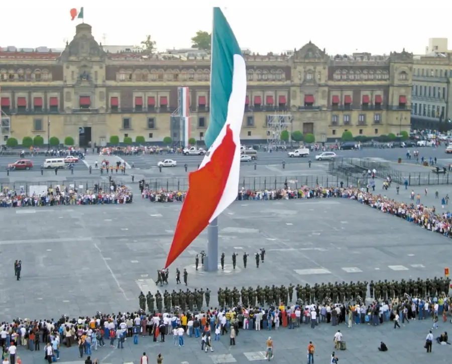 Fotografía del Zócalo de la Ciudad de México con una enorme bandera de México izada al centro, una escolta militar formada y una multitud de personas observando; al fondo se aprecia el Palacio Nacional.