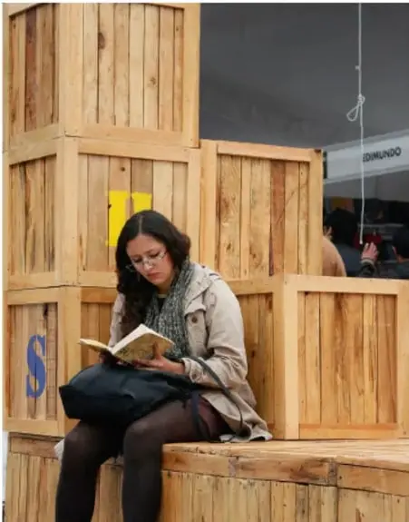 Fotografía de una joven leyendo un libro sentada sobre cajas de madera apiladas, en un espacio público.