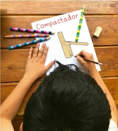 Fotografía de un niño dibujando en una hoja la palabra "Compactador" junto a un boceto de la herramienta, con lápices sobre la mesa.