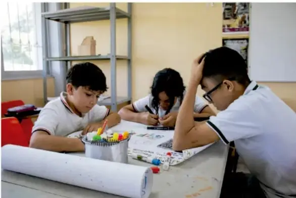 Fotografía de tres estudiantes de telesecundaria sentados en pupitres trabajando juntos en una actividad escrita con marcadores de colores y un rollo de papel sobre la mesa.