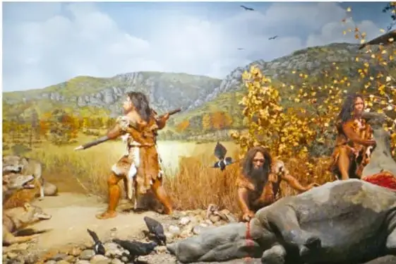 Recreación de un grupo de homínidos prehistóricos cazando junto a un río, con montañas y vegetación al fondo.