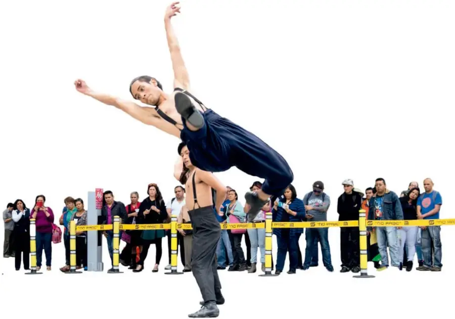 Fotografía de un bailarín realizando un salto acrobático frente a un público diverso, detrás de una cinta amarilla de precaución. Representa la ruptura de estereotipos de género en las profesiones artísticas.