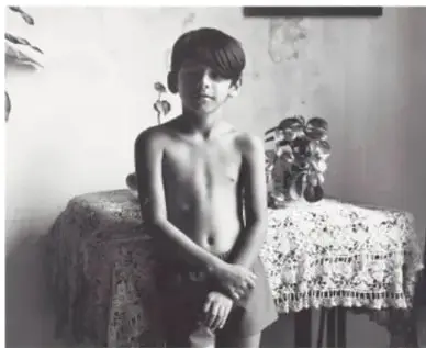 Fotografía en blanco y negro de un niño sin camisa de pie junto a una mesa cubierta con mantel encajado; Niño de Tlacotalpan, 1987, Tlacotalpan, Veracruz, de Mariana Yampolsky (1925-2002).