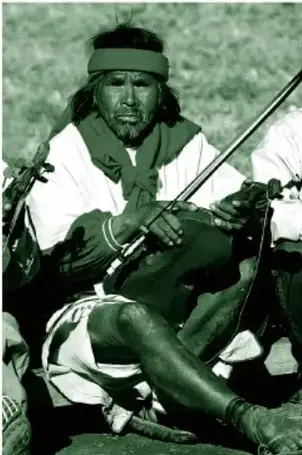 Fotografía a color (en tinta verde del libro) de un hombre ralámuli sentado en el piso con piernas cruzadas, vestido de blanco con distintivos rojos, banda roja al hombro, koyera en la cabeza y huaraches, tocando un violín café con su arco.