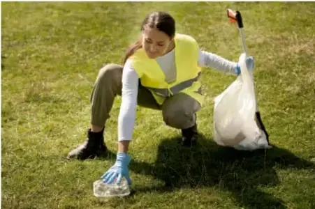 Fotografía de una estudiante con chaleco reflectante y guantes recogiendo residuos (una botella) en un área verde, ejemplo de intervención comunitaria de prevención y cuidado del medio ambiente.