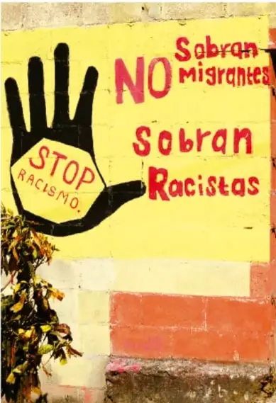 Muro pintado de amarillo con una mano negra y letreros pintados que dicen «Sobran NO migrantes» y «Sobran racistas», con un octágono «STOP racismo»; denuncia graffiti contra la xenofobia.