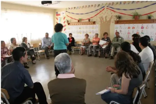 Fotografía de una reunión comunitaria en un salón con adornos, donde un grupo de personas se sienta en círculo en sillas mientras una facilitadora de pie dirige la sesión.
