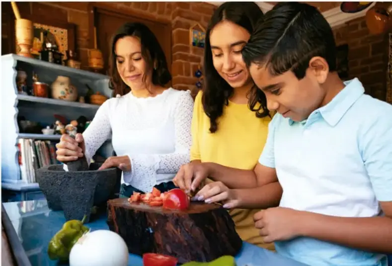 Fotografía de una familia en una cocina: una mujer mayor, una joven y un niño preparan alimentos saludables; hay un molcajete, un cuchillo, jitomate, cebolla y chile.