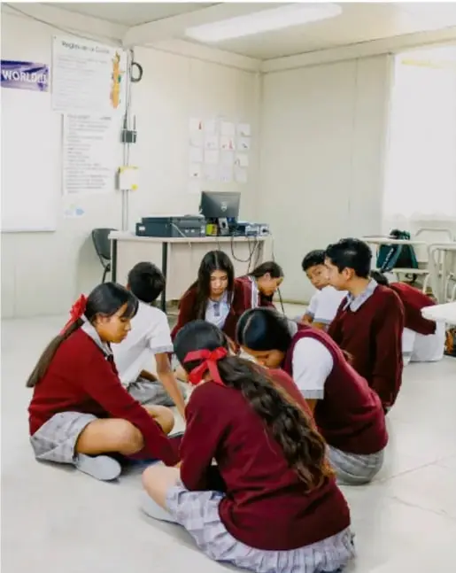 Fotografía de estudiantes de telesecundaria en uniforme rojo, sentados en el piso de un aula trabajando en equipo con materiales escritos.