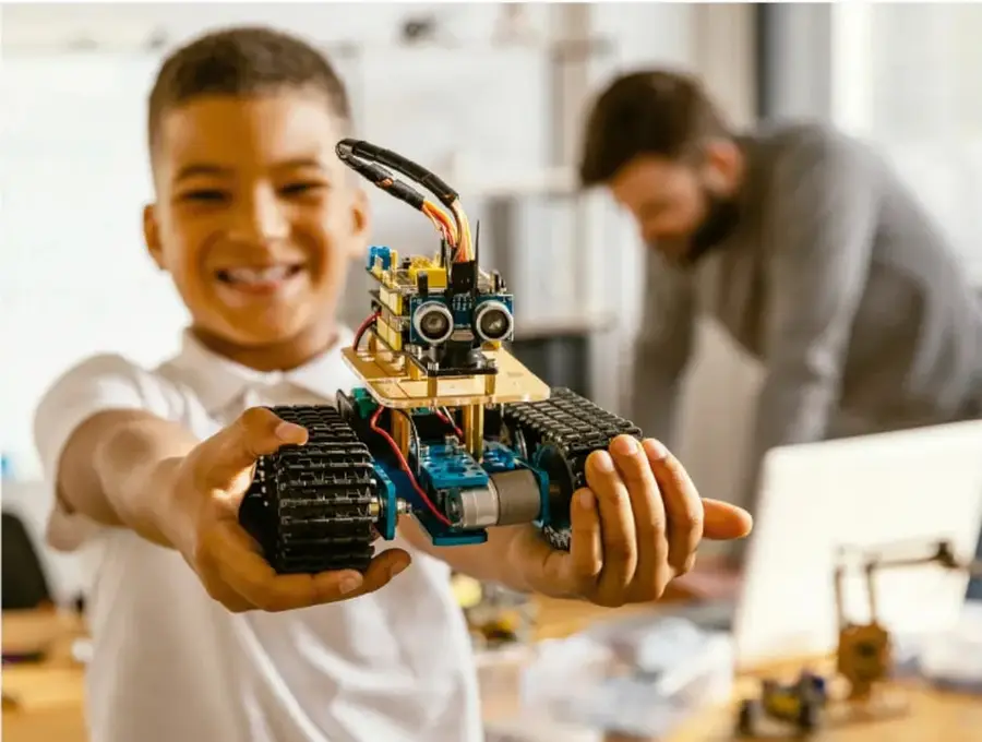 Fotografía a color de un estudiante de secundaria sonriente que extiende los brazos sosteniendo un pequeño robot móvil con orugas de goma, un chasis azul, motores, cables y sensores ultrasónicos montados arriba. Al fondo, desenfocado, otro joven trabaja frente a una computadora en lo que parece ser un taller o laboratorio escolar de robótica.