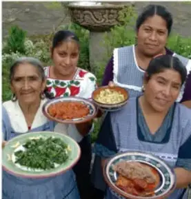 Fotografía de cuatro mujeres indígenas sonrientes sosteniendo platos con platillos típicos preparados, probablemente en un patio o jardín comunitario.