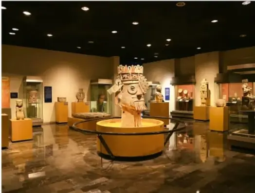 Sala de un museo con vitrinas y una escultura prehispánica iluminada en el centro, probablemente de la Red de Museos del INAH.