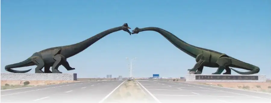 Carretera amplia en el desierto flanqueada por dos monumentos con dinosaurios de metal verde oscuro (sauropodos) de cuello largo que se inclinan hasta tocarse el hocico sobre la vía; frontera entre China y Mongolia en Erenhot.