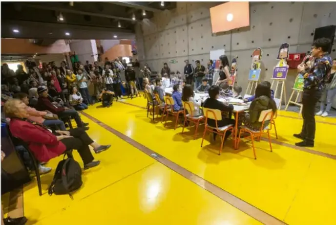 Galería escolar en un patio con público sentado, mesas de exposición y una persona hablando por micrófono.