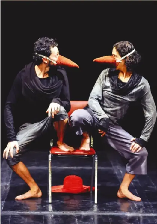 Dos personas actúan en un escenario oscuro, vestidas de gris y negro, con narices largas rojizas de personaje; una apoya el pie en una silla roja y en el suelo hay un sombrero rojo.