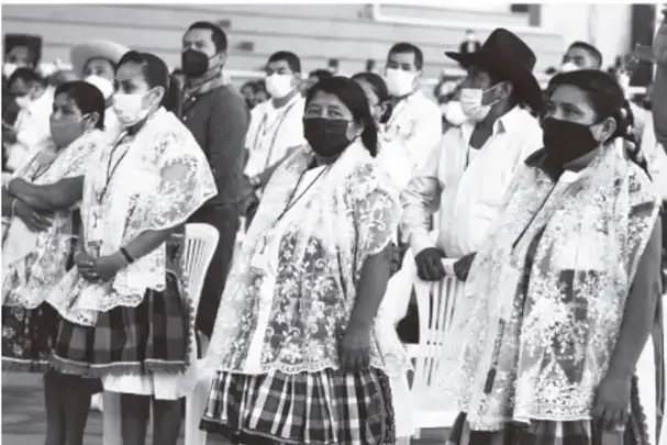 Fotografía en blanco y negro de mujeres indígenas con vestimenta tradicional (quechquémetl bordado, faldas de cuadros, cubrebocas) reunidas en un acto público; al fondo varios hombres, uno con sombrero.