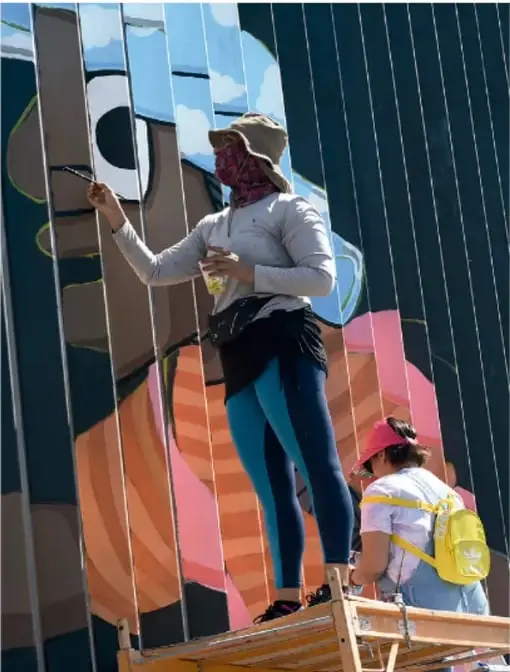 Fotografía de una artista con sombrero, mascada y mascarilla pintando un mural colorido en una estructura metálica, mientras otra persona con mochila amarilla observa desde abajo; representa el arte callejero o muralismo.