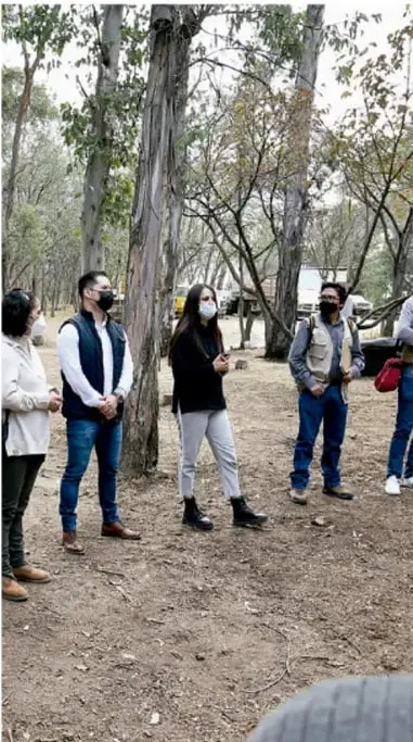 Fotografía de un grupo de personas (estudiantes y docentes) con cubrebocas, de pie en un espacio al aire libre con árboles, conversando en círculo.