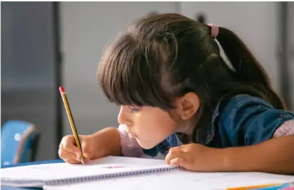 Fotografía de una niña de primaria sentada en su escritorio, concentrada escribiendo en su cuaderno con un lápiz, representando la actividad de responder las preguntas en comunidad.
