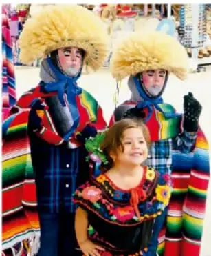Dos personas disfrazadas con vestimenta tradicional colorida y sombreros de pelaje, junto a una niña en traje típico, en una celebración comunitaria.