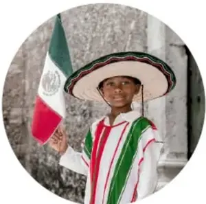 Fotografía circular de un niño sonriente con traje tradicional de manta bordada en verde y rojo, sombrero de palma con listón tricolor y una bandera de México en la mano, frente a un muro de piedra.