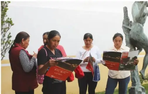 Grupo de personas, en su mayoría mujeres, revisa folletos informativos al aire libre; al fondo se observa una escultura.