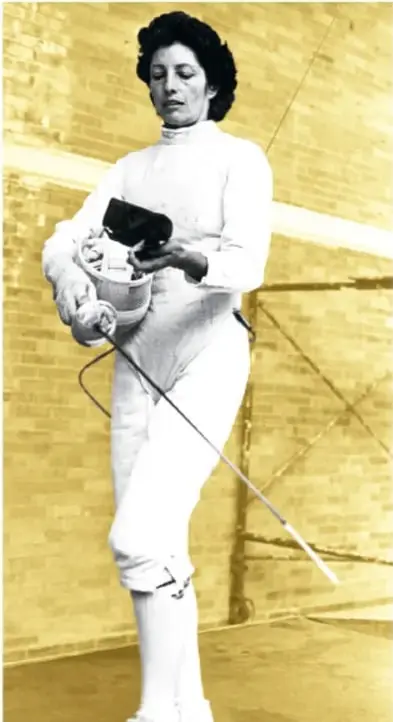 Fotografía histórica con tinte amarillo de una esgrimista con traje blanco completo, sosteniendo en un brazo la máscara de esgrima y en la mano un florete (espada de esgrima), de pie en una instalación deportiva.