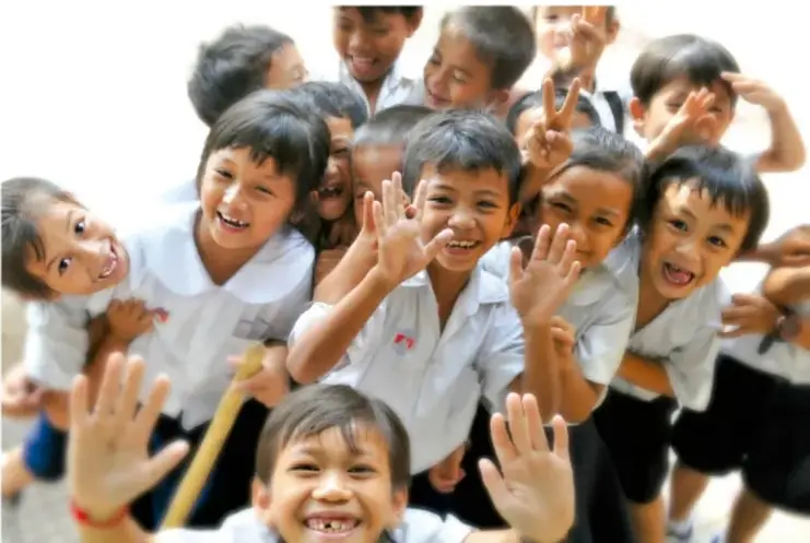 Fotografía de un grupo de niñas y niños sonrientes, con uniforme escolar, levantando las manos y haciendo señas de paz; reflejan alegría y diversidad.
