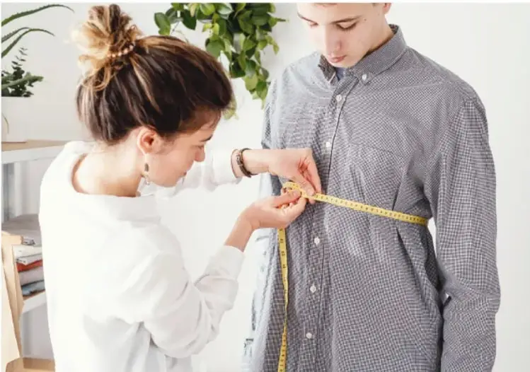 Fotografía de una joven tomando medidas con una cinta métrica amarilla alrededor del torso de un joven con camisa de cuadros, ilustrando el uso de unidades básicas de longitud en la vida cotidiana.