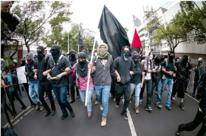 Fotografía de una marcha o protesta ciudadana: grupo de personas con el rostro cubierto caminando por una avenida, portando banderas negras y rojas, en un entorno urbano con edificios y árboles.