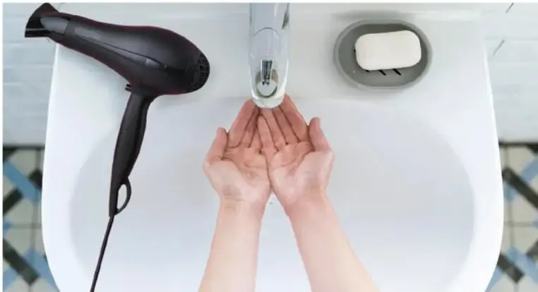 Fotografía de una persona lavándose las manos en un lavabo con una secadora de cabello conectada cerca, ilustrando el riesgo de usar aparatos eléctricos en el baño.