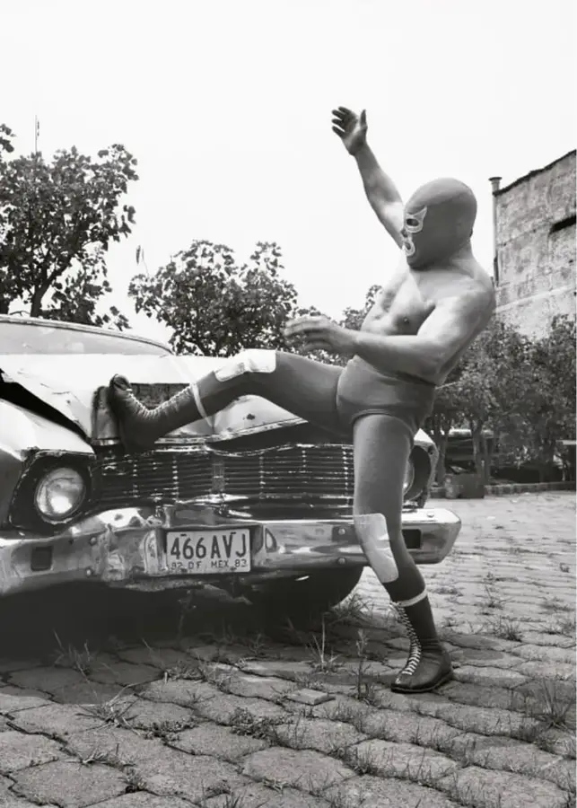 Fotografía en blanco y negro de Blue Demon, luchador enmascarado, con el pie apoyado sobre el cofre de un automóvil antiguo, en los Estudios Churubusco, 1984.
