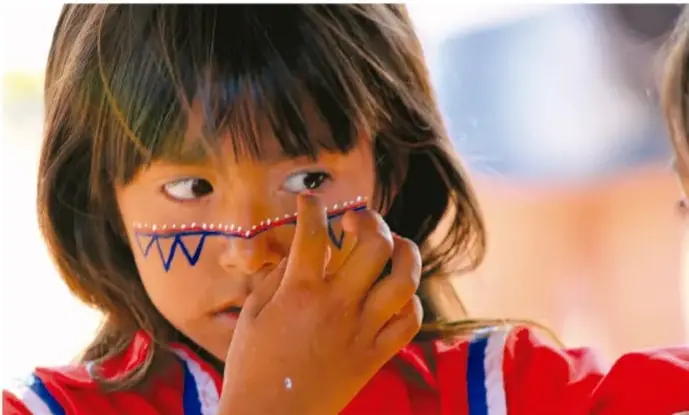 Fotografía a color de una niña indígena con pelo castaño y blusa roja con detalles azul y blanco, que se pinta una línea punteada azul con dibujos zigzag sobre la mejilla, expresión seria y mirada lateral.