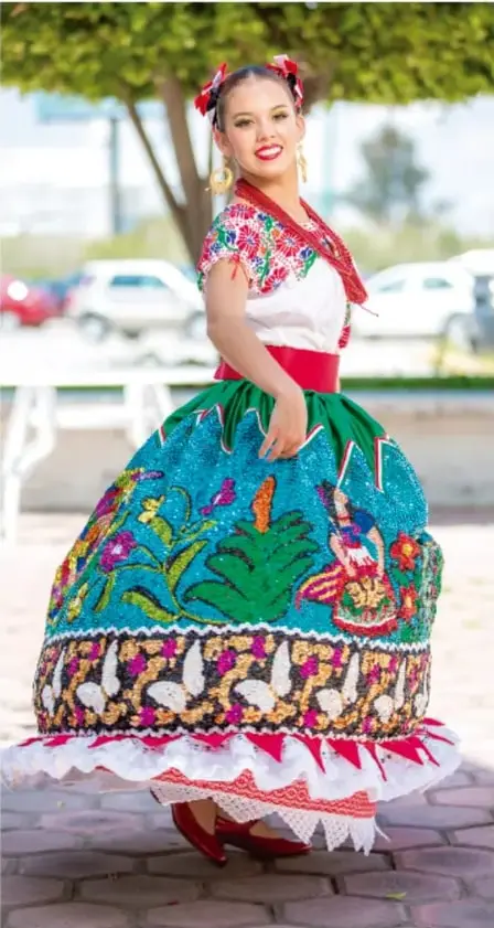 Fotografía a color de una joven posando con un traje típico mexicano: falda amplia turquesa con bordados de flores, aves y follaje en rojo, verde y amarillo, enaguas con volados de encaje blanco, blusa blanca con bordados en el cuello, cinta roja en la cintura y moños rojos en el cabello. Está parada sobre adoquines, con árboles y autos al fondo.