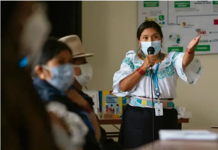 Mujer indígena con mascarilla y blusa bordada, hablando con micrófono ante un grupo de personas también con mascarillas, en un acto comunitario de promoción de la salud; fondo con carteles informativos.