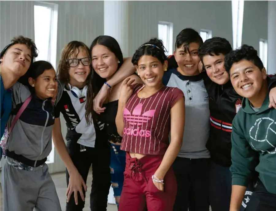 Fotografía de un grupo de ocho adolescentes de secundaria, sonrientes, abrazados y apoyados unos sobre otros en un pasillo iluminado, representando la amistad y el compañerismo.