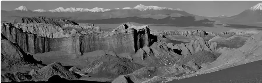 Paisaje desértico del Atacama con formaciones rocosas erosionadas, cerros y volcanes nevados al fondo.