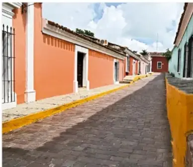 Calle empedrada con casas de colores, árboles y cielo nublado.