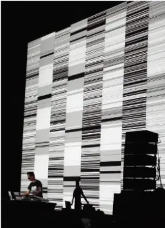 Fotografía en blanco y negro de una instalación sonora: una gran pantalla o muro con patrones de líneas horizontales, con figuras humanas y equipo de audio al pie.