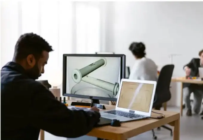 Fotografía de un oficina o taller de diseño: un trabajador observa en una pantalla de escritorio un modelo 3D de una pieza mecánica, con un portátil abierto sobre la mesa; al fondo otras personas trabajan en sus estaciones.