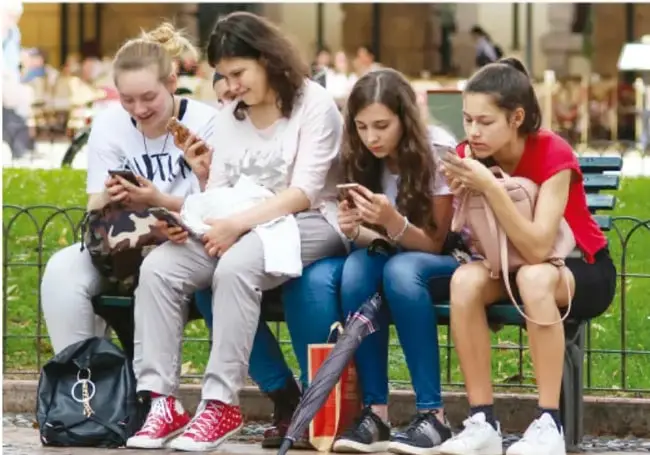 Cuatro jóvenes sentadas en una banca en un parque, todas utilizando sus teléfonos celulares al mismo tiempo, ilustrando el uso cotidiano de los dispositivos móviles y redes sociales.