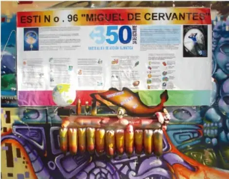 Fotografía de un periódico mural escolar titulado 'ESC. N.o. 96 Miguel de Cervantes' conmemorando los 500 años de su muerte, montado sobre un muro con graffiti colorido.