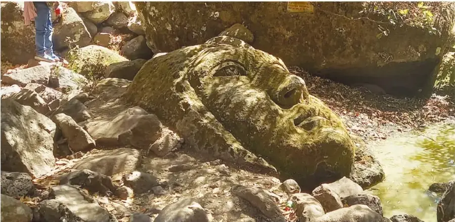 Fotografía panorámica de una cabeza gigante recostada entre piedras junto a un arroyo de agua clara; una persona con pantalón azul y camiseta rosa la observa desde las rocas.