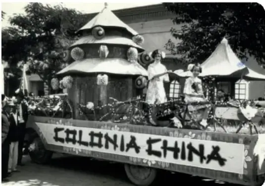 Fotografía en blanco y negro de un desfile con una carroza alegórica que tiene un letrero que dice 'COLONIA CHINA'. Se observan personas y una estructura decorada.