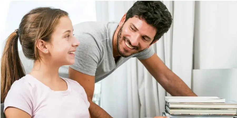 Fotografía de un joven (estudiante) y un adulto varón sonriendo y conversando junto a una pila de libros, en un ambiente luminoso que ilustra el diálogo intergeneracional.